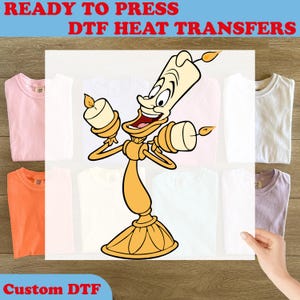 May include: A DTF heat transfer featuring a cartoon illustration of a golden candelabra character with a smiling face and lit candles on its arms. The text READY TO PRESS DTF HEAT TRANSFERS and Custom DTF appear in red against a wooden background.