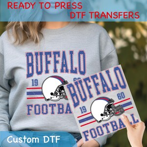 May include: Gray sweatshirt and DTF transfer featuring "BUFFALO FOOTBALL" in blue and red, with a football helmet graphic. The transfer is held up to the sweatshirt, showing the design. Text on the image reads "READY TO PRESS DTF TRANSFERS" and "Custom DTF".