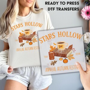 May include: A cream-colored t-shirt with the words "STARS HOLLOW" in orange, along with an illustration of a coffee cup, book, candle, and pumpkins. The text "ANNUAL AUTUMN FESTIVAL" is also present. A DTF transfer with the same design is held up.