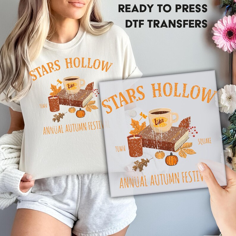 May include: A cream-colored t-shirt with the words "STARS HOLLOW" in orange, along with an illustration of a coffee cup, book, candle, and pumpkins. The text "ANNUAL AUTUMN FESTIVAL" is also present. A DTF transfer with the same design is held up.