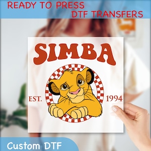 May include: A white square DTF transfer featuring the word "SIMBA" in orange retro font above a cartoon lion cub. The lion cub is inside a checkered circle with "EST. 1994" below. The words "READY TO PRESS DTF TRANSFERS" and "Custom DTF" are also present.