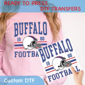 May include: A light pink sweatshirt with a distressed blue and red "BUFFALO FOOTBALL" graphic, featuring a football helmet. A custom DTF transfer with the same design is held in front. Text at the top reads "READY TO PRESS DTF TRANSFERS".