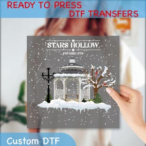 May include: A gray DTF transfer featuring a winter scene of Stars Hollow, with a gazebo, snow, and a tree with red birds. The text "STARS HOLLOW FOUNDED 1779" is at the top. The words "READY TO PRESS DTF TRANSFERS" and "Custom DTF" are also visible.