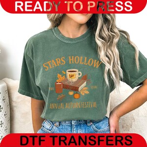 May include: Green t-shirt with the text "STARS HOLLOW ANNUAL AUTUMN FESTIVAL" in orange. The design includes a coffee cup, books, leaves, a candle, and a pumpkin. The shirt is worn by a person.
