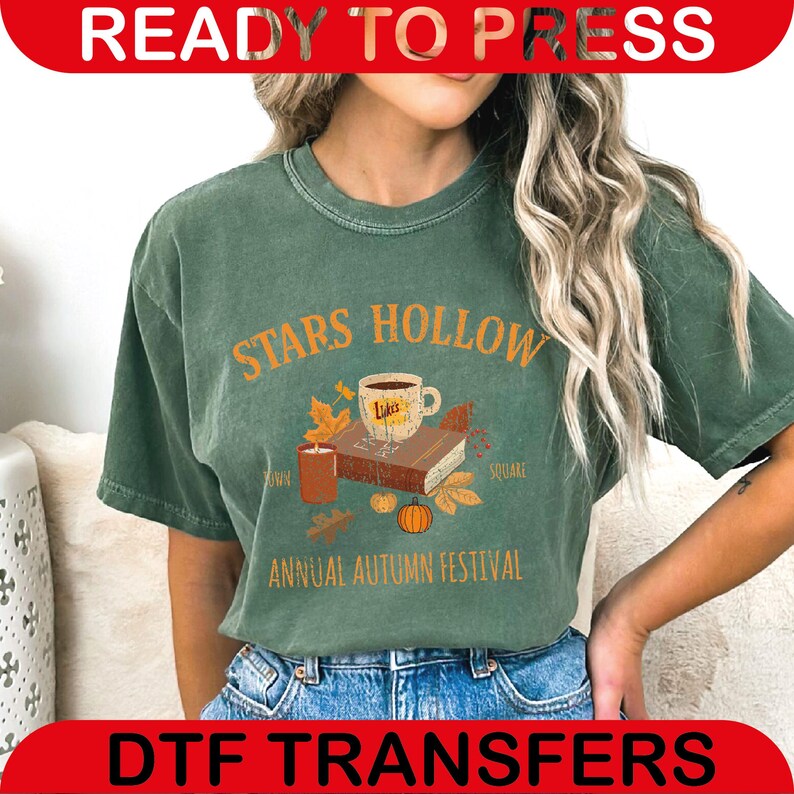 May include: Green t-shirt with the text "STARS HOLLOW ANNUAL AUTUMN FESTIVAL" in orange. The design includes a coffee cup, books, leaves, a candle, and a pumpkin. The shirt is worn by a person.