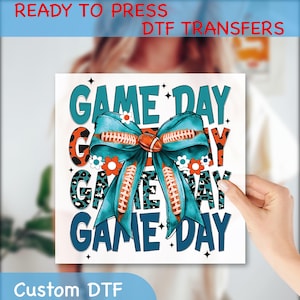May include: A white DTF transfer with the words "GAME DAY" in teal and black, featuring a football-shaped bow with football laces. The transfer also includes the text "READY TO PRESS DTF TRANSFERS" and "Custom DTF".