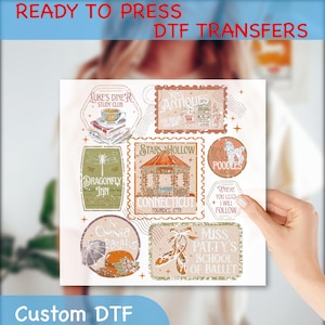 May include: A sheet of ready-to-press DTF transfers featuring various designs. Designs include a diner, antique shop, gazebo, ballet shoes, and other vintage-style graphics. The sheet is held by a person's hand.