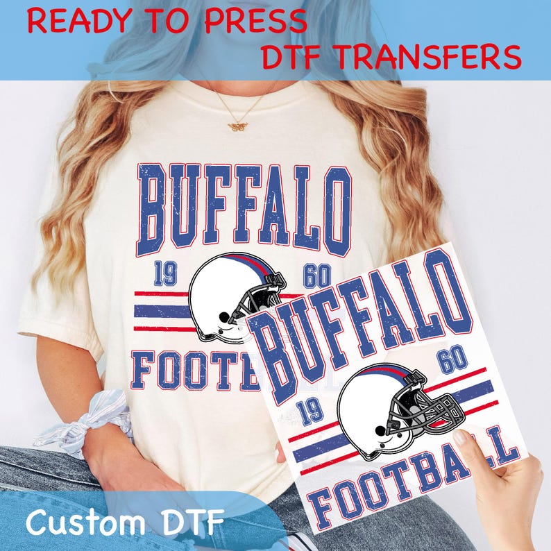 May include: Cream-colored t-shirt with a distressed blue "BUFFALO FOOTBALL" graphic, featuring a football helmet and the numbers "19" and "60". A DTF transfer with the same design is held in front. Text at the top reads "READY TO PRESS DTF TRANSFERS".