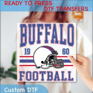 May include: A white square DTF transfer featuring the word "BUFFALO" in blue and red distressed text, with a football helmet graphic. The transfer also includes the words "FOOTBALL", "19", and "60". The text "READY TO PRESS DTF TRANSFERS" and "Custom DTF" are also present.