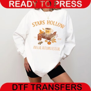 May include: White sweatshirt with a graphic featuring the text "STARS HOLLOW ANNUAL AUTUMN FESTIVAL". The design includes a coffee cup, book, leaves, and a pumpkin. The words "READY TO PRESS" and "DTF TRANSFERS" are also visible.