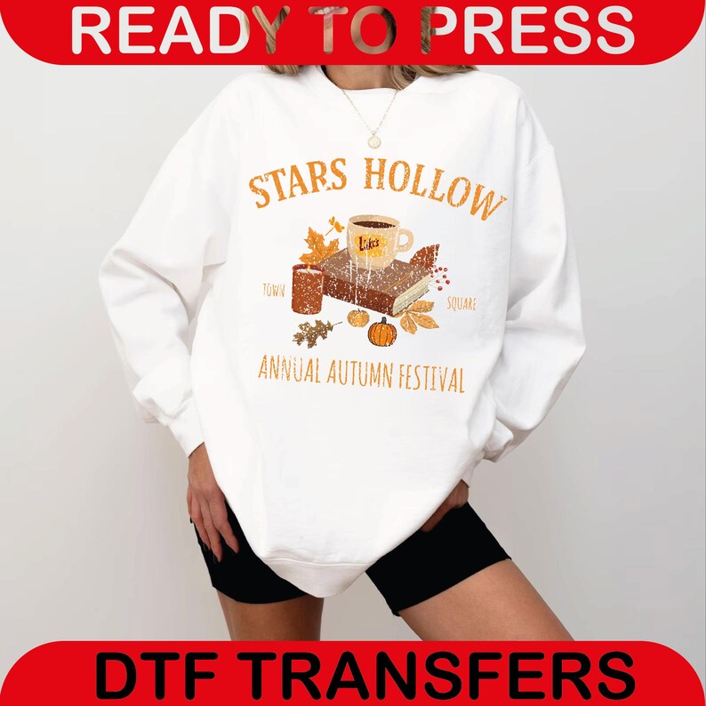 May include: White sweatshirt with a graphic featuring the text "STARS HOLLOW ANNUAL AUTUMN FESTIVAL". The design includes a coffee cup, book, leaves, and a pumpkin. The words "READY TO PRESS" and "DTF TRANSFERS" are also visible.
