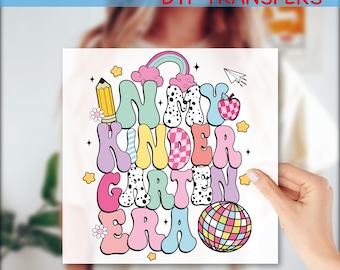 May include: A colorful DTF transfer design with the text "In My Kindergarten Era." The design features a rainbow, apple, pencil, and disco ball. The words are in bubble letters with various patterns and colors, perfect for a school-themed project.