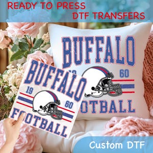 May include: White pillow and DTF transfer featuring a distressed design with the word "BUFFALO" in blue, a football helmet, and the word "FOOTBALL". The text "READY TO PRESS DTF TRANSFERS" and "Custom DTF" are also visible.