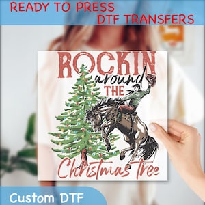 May include: A white DTF transfer featuring a cowboy riding a bucking horse next to a Christmas tree. The text reads "Rockin' around the Christmas Tree". The transfer is held by a person.