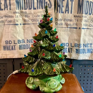May include: A glossy green ceramic Christmas tree with a textured, tiered design and a star-shaped top. The tree is decorated with small, multi-colored plastic light bulbs in red, blue, orange, and purple, and features a white electrical cord.