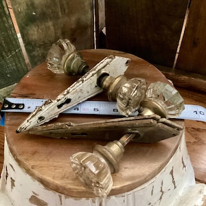 May include: Antique door hardware set featuring clear glass knobs and aged brass accents. Includes a white painted door plate and a measuring tape. The hardware is likely from the early 20th century.