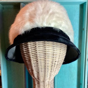 May include: A white fur hat with a black velvet brim. The hat is on a wicker hat form.