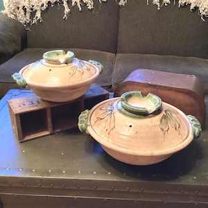 May include: Two ceramic cooking pots with lids. The pots are light pink with green accents and leaf designs. The lids have a small hole in the center. The pots are sitting on a green metal surface.