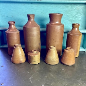 May include: A collection of seven brown stoneware bottles of various sizes and shapes. The bottles have a rustic, vintage look and are perfect for displaying or using as decorative accents.
