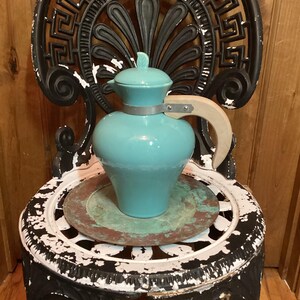 May include: A turquoise ceramic pitcher with a matching lid featuring a small leaf-shaped handle. The pitcher has a curved light-colored wooden handle attached with a silver-toned metal band that encircles the neck of the vessel.