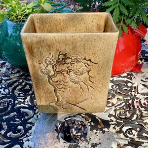 May include: A square, tan ceramic planter with a tree design. The planter has a textured surface and a light brown color with darker brown accents. The tree design is in a darker brown color. The planter is sitting on a decorative metal table.
