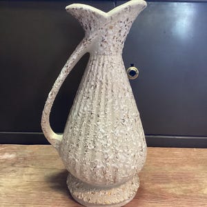 May include: A vintage white ceramic pitcher with a textured surface and gold flecks. The pitcher has a handle and a flared spout. The base is round and also has gold accents. The pitcher is likely used for decorative purposes.