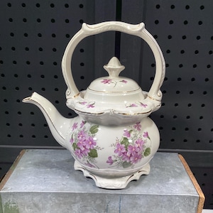May include: A cream-colored ceramic teapot featuring a floral pattern of purple violets and green leaves. The teapot has a scalloped base, a matching lid with a pointed finial, and a high arched handle, all accented with thin gold trim.