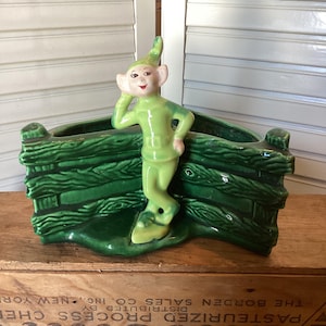 May include: A vintage ceramic planter featuring a green elf figure leaning against a green fence-like structure. The elf wears a green outfit and a pointed hat. The planter is set on a wooden crate.