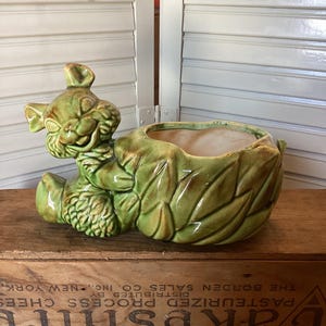 May include: A vintage green ceramic planter shaped like a smiling bear holding a leaf-shaped pot. The planter has a glossy finish and is sitting on a wooden crate. The crate has text that reads "Lakeshire Pasteurized Process Cheeses".