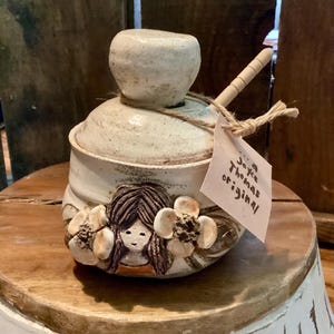 May include: A handmade ceramic jar with a lid and a small wooden spoon. The jar is off-white with a textured surface and features a sculpted face surrounded by flowers. A tag reads "Joyce Thomas original."