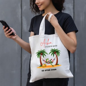 May include: White tote bag with the text "Acho, Puerto Rico" in red script. The bag features a cartoon frog playing a guitar in a hammock between two palm trees. The phrase "Es Otra Cosa" is printed below.