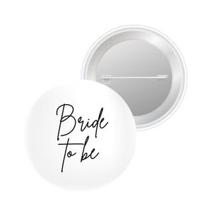 May include: White button with the text "Bride to be" in black script. The pin is round and has a safety pin on the back. The second button is shown from the back, with a silver metal finish.