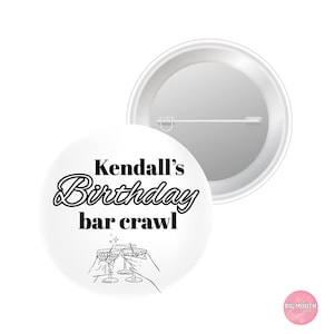May include: A round white pin-back button featuring the black text Kendall's Birthday bar crawl above a line drawing of three hands holding cocktail glasses. The back of the button is silver-toned metal with a horizontal pin mechanism attached.