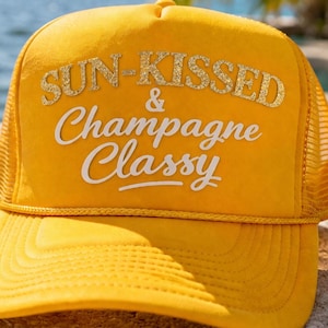 May include: A yellow trucker hat with the text "SUN-KISSED & Champagne Classy" in gold glitter and white lettering. The hat has a mesh back and a curved brim. Sunglasses and a glass of champagne are in the background.