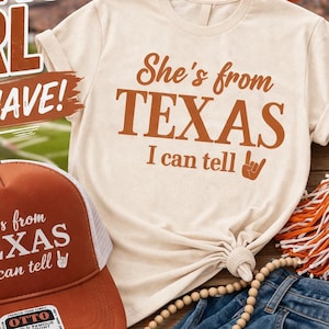 May include: A cream-coloured t-shirt and a trucker hat with the text "She's from TEXAS I can tell" in brown. The hat is orange with a white mesh back. The image also includes a football, denim shorts, and a Texas-shaped charm.