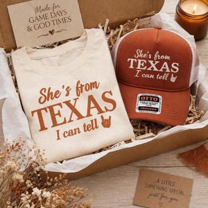 May include: A gift box with a cream t-shirt and a burnt orange trucker hat, both with the text "She's from TEXAS I can tell". The box also contains a small candle and a card that reads "A little something special just for you". The box is filled with paper.