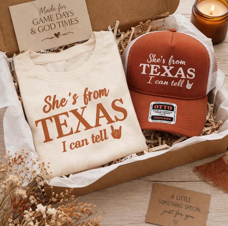 May include: A gift box with a cream t-shirt and a burnt orange trucker hat, both with the text "She's from TEXAS I can tell". The box also contains a small candle and a card that reads "A little something special just for you". The box is filled with paper.