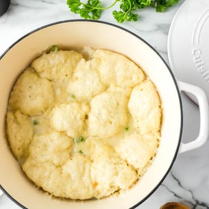 May include: Overhead shot of a creamy chicken and dumpling dish in a cream-colored pot. The dumplings are golden brown and sit atop a creamy sauce with visible peas. A white lid, parsley, and a wooden spoon are nearby.