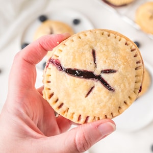 May include: A hand holding a golden-brown, round blueberry pie with a star-shaped vent. The pie has a crimped edge and a dark purple filling. Other pies and blueberries are in the background.