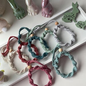 Op de afbeelding: Een verzameling kleurrijke macramé armbanden met houten kralen, gepresenteerd op een witte tray samen met keramische konijnenbeeldjes en houten konijnenuitsparingen. De armbanden zijn in roze, blauw, groen en grijs.