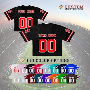 May include: A black V-neck jersey with red and white trim on the sleeves, featuring the text "TEAM NAME" and the number "00" in red with white outlines. The back displays "YOUR NAME" and "00". Includes a palette icon and "CUSTOM ANY DESIGN".