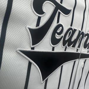 May include: White baseball jersey with black vertical stripes and a black "Team" patch on the front.