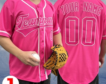 Pode incluir: Camisola de beisebol rosa com guarnições brancas e um gráfico branco de "Teammates" na frente. A parte de trás da camisola tem um "YOUR NAME" branco e um número branco "00".