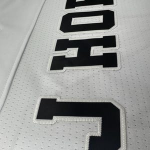 Pu&ograve; includere: Maglia bianca con lettere nere cucite che compongono la scritta "JONES" in un carattere grande.