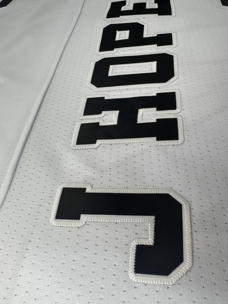 Pu&ograve; includere: Maglia bianca con lettere nere cucite che compongono la scritta "JONES" in un carattere grande.