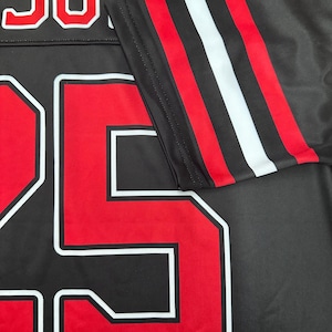 May include: Black jersey with the name "JON" in white-outlined red letters at the top. The number "25" is prominently displayed in red with a white outline. The sleeve features red, white, and black stripes.