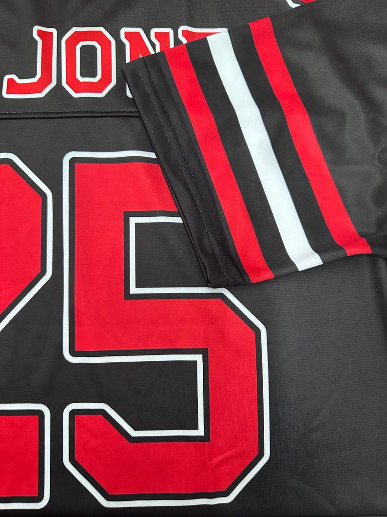 May include: Black jersey with the name "JON" in white-outlined red letters at the top. The number "25" is prominently displayed in red with a white outline. The sleeve features red, white, and black stripes.