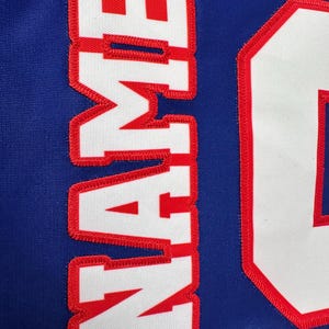 May include: White jersey with red stitching, the word "NAME" is sewn on the front in white letters with red outlines.