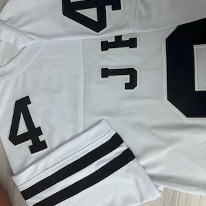 Pu&ograve; includere: Maglia bianca con doppie strisce nere sulle maniche. La maglia ha un numero nero "4" sul davanti e un numero nero "4" sulla manica sinistra. La maglia ha anche una lettera nera "J" e un numero nero "4" sul retro.