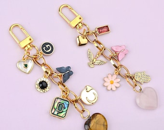 May include: Two gold-coloured keychains with various charms. One keychain has a heart with a green gem, a black and gold charm, and a square abalone charm. The other has a pink butterfly, a gold bird, and a pink flower charm.